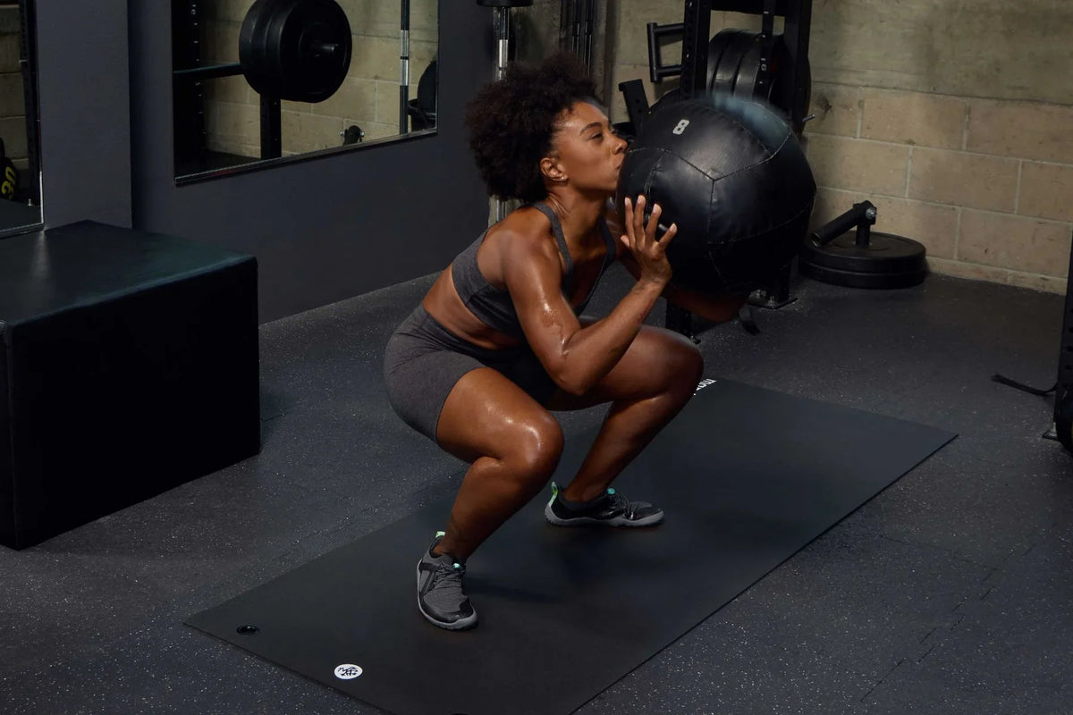 Woman in a gym, squatting, holding a weighted ball, on a Manduka P/ROX Hybrid Fitness Mat | Eco Yoga Store