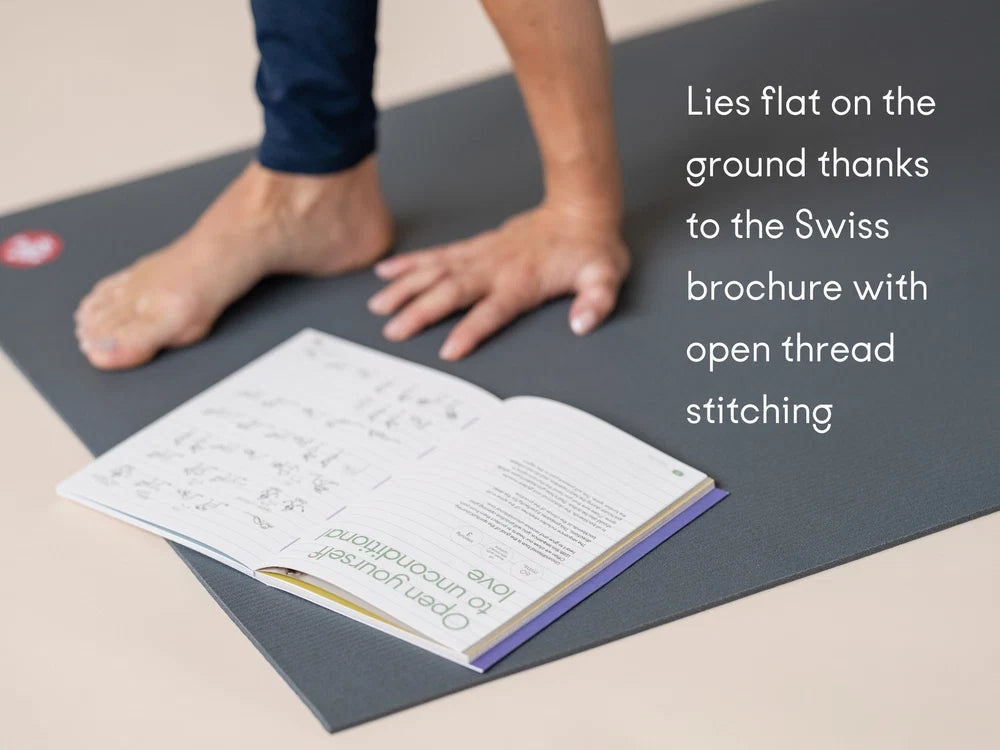 A Manduka PROLite yoga mat on a beige floor with a person's bare foot and hand visible, alongside an open Chakra Flow book. White text on the right reads: 'Lies flat on the ground thanks to the Swiss brochure with open thread stitching' - Yoga Journaling | Eco Yoga Store