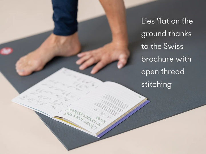 A Manduka PROLite yoga mat on a beige floor with a person's bare foot and hand visible, alongside an open Chakra Flow book. White text on the right reads: 'Lies flat on the ground thanks to the Swiss brochure with open thread stitching' - Yoga Journaling | Eco Yoga Store