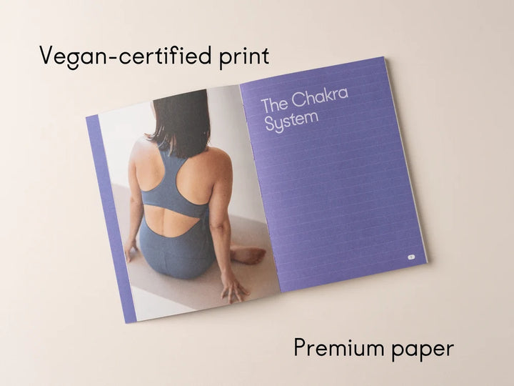 An open Chakra Flow book laid on a light beige background, showing a purple page with the title 'The Chakra System' and a printed photo of a seated person. Text above reads 'Vegan-certified print' and below reads 'Premium paper' - Yoga Journaling | Eco Yoga Store