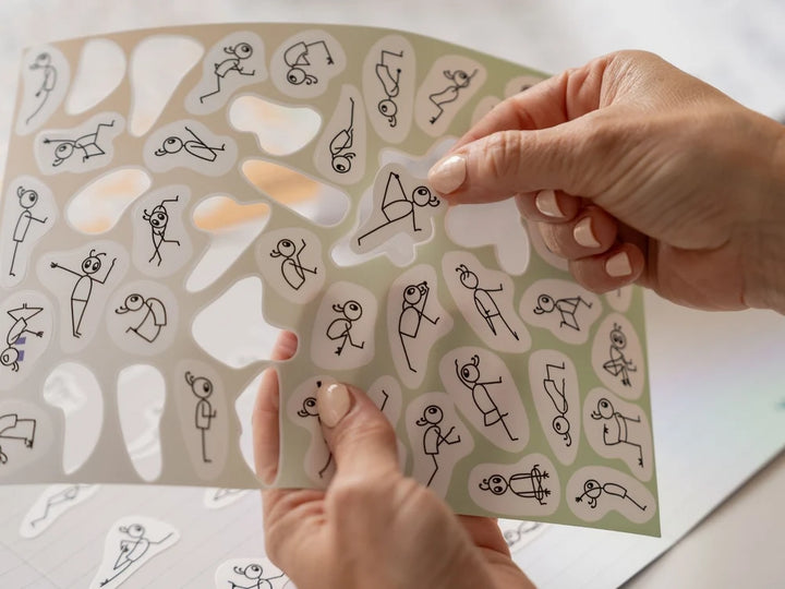 A close-up of hands holding a clear sheet of small die-cut Stick Play Flow stickers printed with simple black line drawings of stick figures in various poses, over a light neutral background - Yoga Journaling | Eco Yoga Store