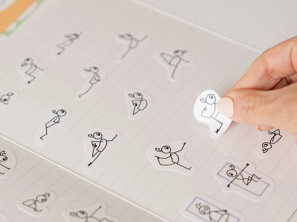 A close-up of a hand placing a Stick Play Flow sticker with a black line drawing of a person in a yoga pose on a lined notebook page, with other similar stickers scattered around - Yoga Journaling | Eco Yoga Store