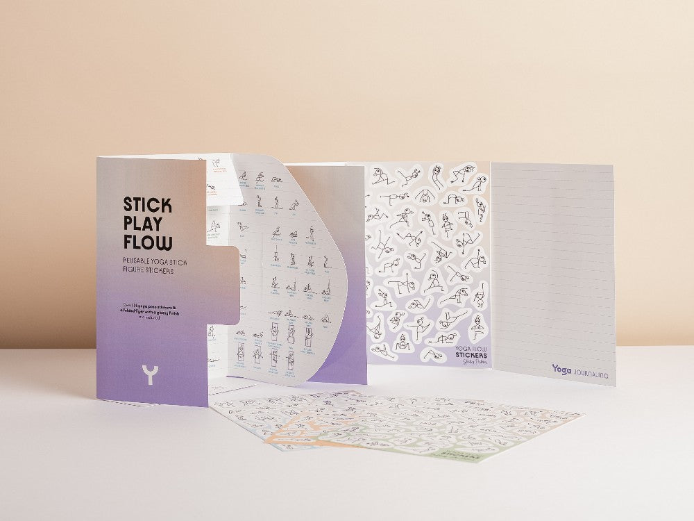 A yoga flow sticker kit displayed on a white surface against a warm beige background. The kit includes a purple and white envelope labeled 'STICK PLAY FLOW', a sheet of small black-and-white yoga pose stickers, a lined practice journal, and a flat illustrated guide sheet - Yoga Journaling | Eco Yoga Store