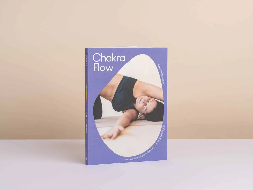 Chakra Flow book standing upright on a white surface against a beige background. The book cover is lavender purple with a large white oval containing a photo of a woman in a navy sports bra performing a yoga pose - Yoga Journaling | Eco Yoga Store