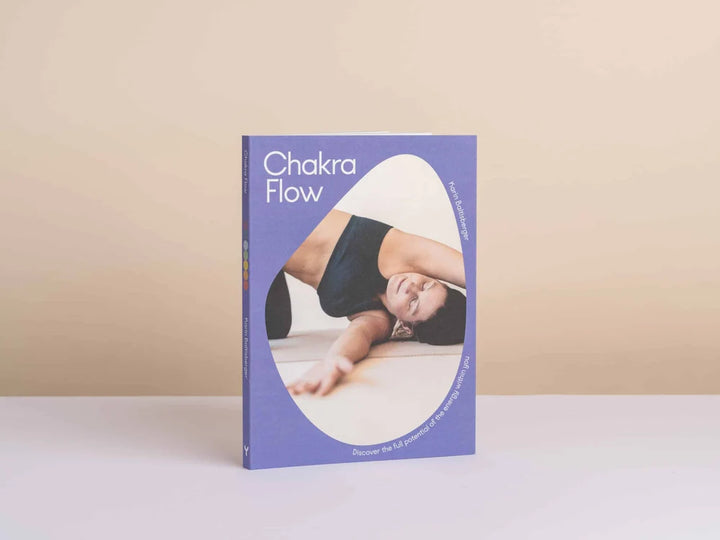 Chakra Flow book standing upright on a white surface against a beige background. The book cover is lavender purple with a large white oval containing a photo of a woman in a navy sports bra performing a yoga pose - Yoga Journaling | Eco Yoga Store