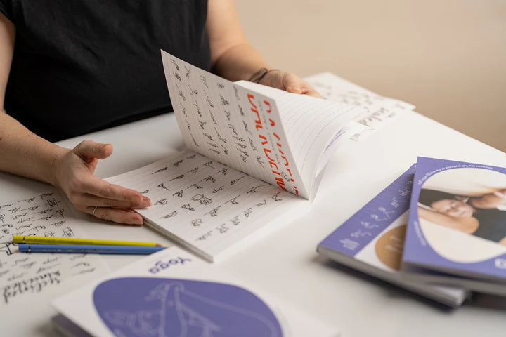 A person's hands holding and flipping through a Yoga Journaling Notebook with handwritten calligraphy. On the table are two colored pencils and a stack of Yoga Journaling Notebooks books - Yoga Journaling | Eco Yoga Store